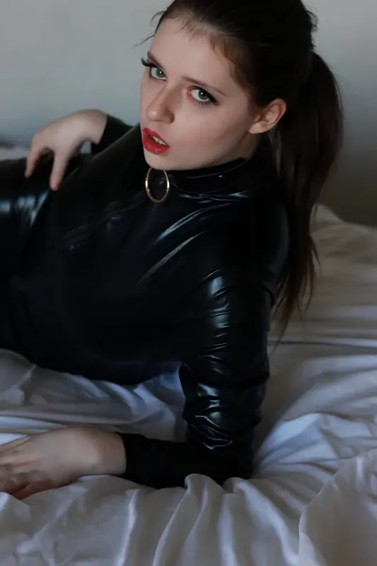 Em Ambisious in a black latex catsuit on a bed