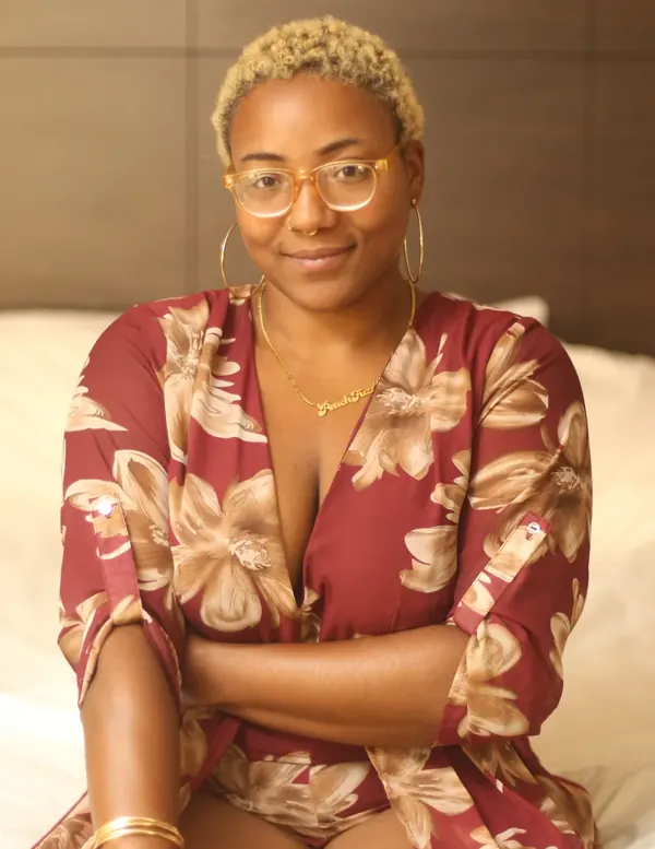 Peach Fuzz smiling in round glasses and a red floral wrap dress, seated on a bed