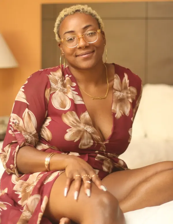Peach Fuzz in a red floral dress and round glasses, sitting on a bed with a soft smile