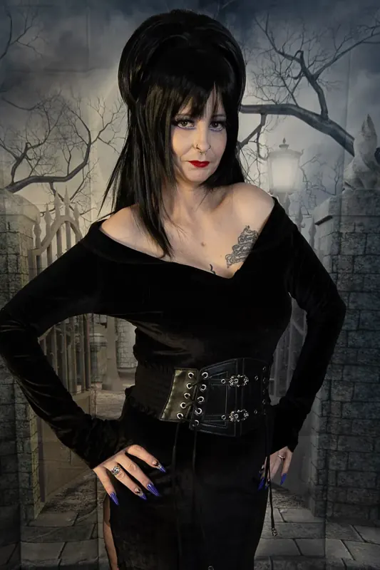 Selina Dark in a black off-shoulder gown and corset belt in front of a gothic backdrop